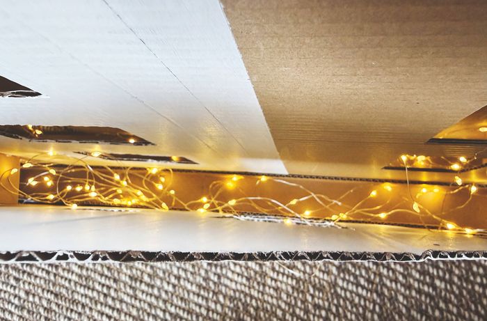 A hand using a wooden peg to attach a red-painted craft star to a string of fairy lights in front of a window.] 