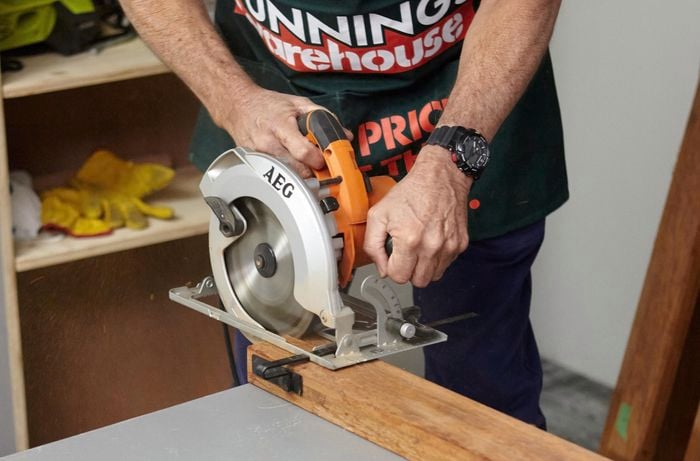 Person using circular saw to cut piece of timber into two pieces.