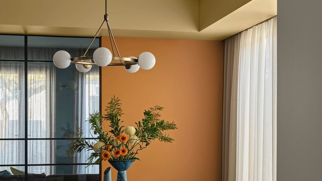 A feature wall painted in Dulux Magic Melon, with windows draped by curtains, a feature pendant light and a vase of flowers in the same colour scheme.