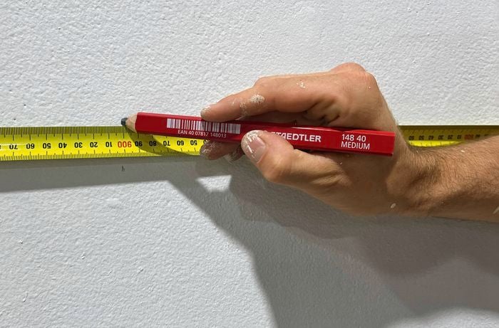  A hand right with paint splatters holds a lead pencil and marks a white wall using a yellow measuring tape.