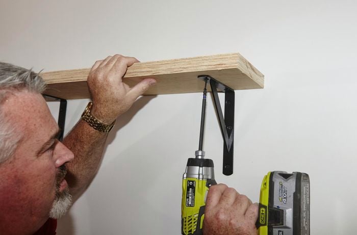 A person screwing a bracket to a timber shelf from underneath using a cordless drill