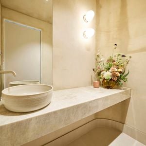 A modern bathroom with a sink on a benchtop with a vase with flowers in it on the benchtop, a mirror and lights on the wall.