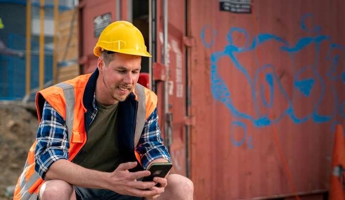 Construction worker in high-visibility vest and yellow hard hat sitting beside a shipping container, using a smartphone, with another worker and scaffolding in the background.
