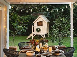 An outdoor seating area with a dining table, a children's cubby house in the garden and hanging festive light bulbs.
