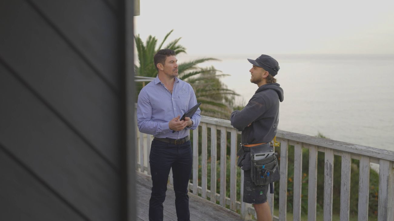 Two men, one in a blue and white stripped shirt holding a tablet and another in a hat, hoodie and tool belt, stand on a balcony outdoors with the ocean as their background]
