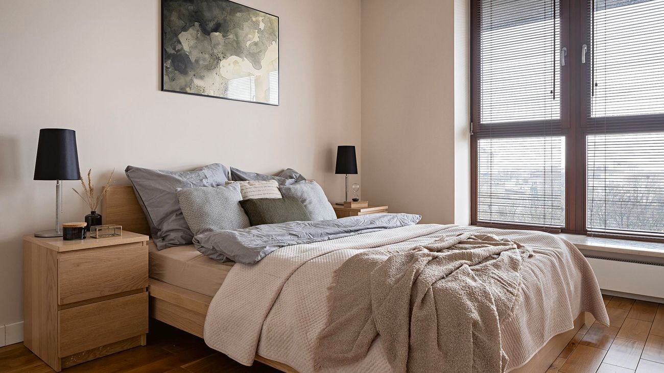 Neutral-coloured bedroom with light wooden bed frame and bedside tables, with a window covered by venetian blinds.