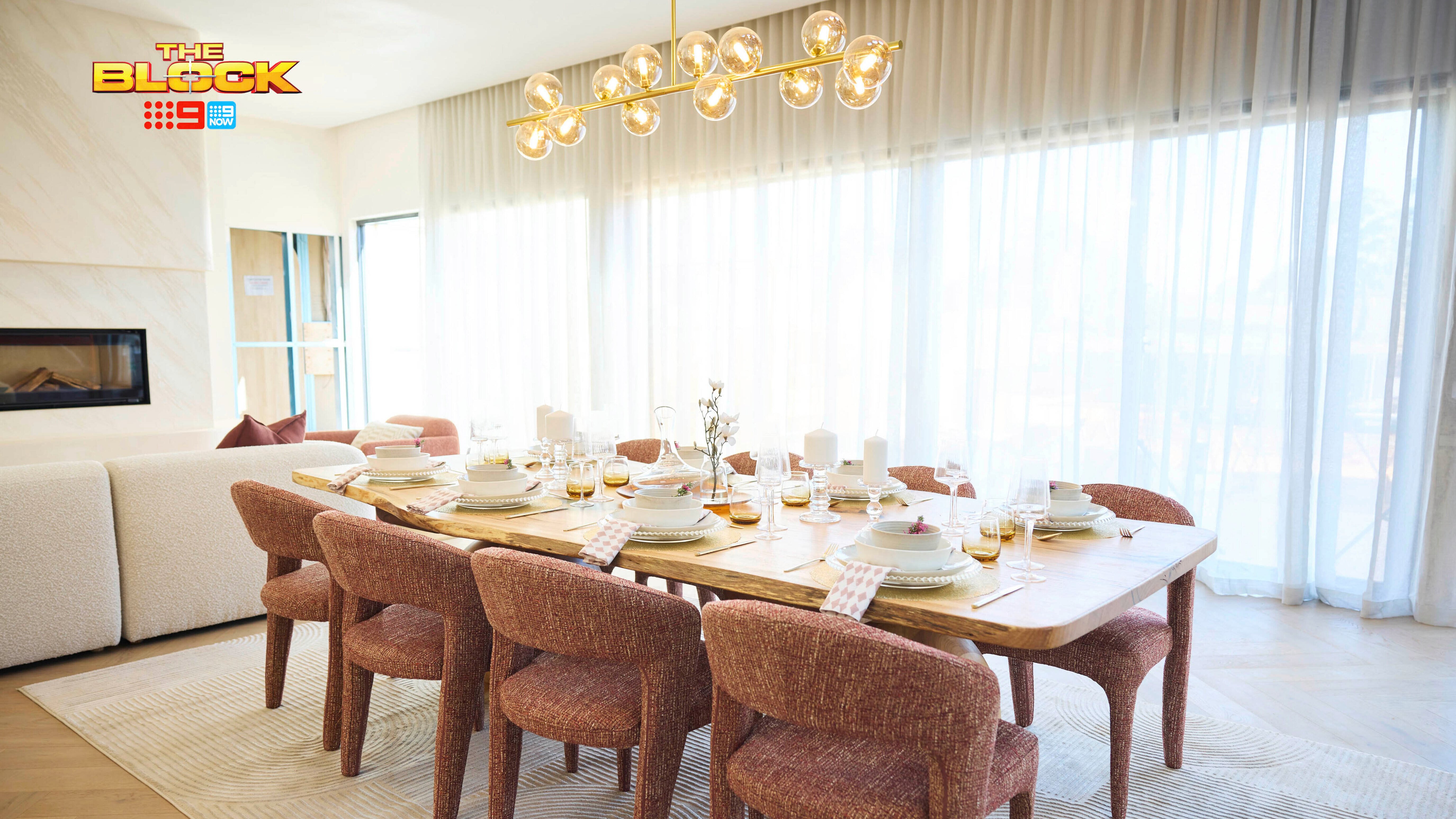 A well-decorated dining room featuring a large wooden table set with plates, bowls, glasses, and napkins, surrounded by eight reddish-brown upholstered chairs. An elegant chandelier with spherical bulbs hangs above. Large windows with sheer white curtains let in natural light. A white sofa and part of another room are visible through an open doorway.