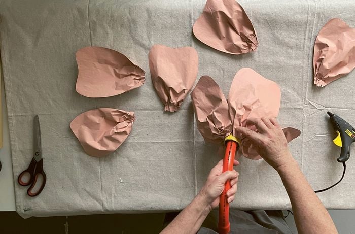 Hands assembling peach-coloured paper petals onto an orange conduit bend, with a hot-glue gun and scissors nearby.