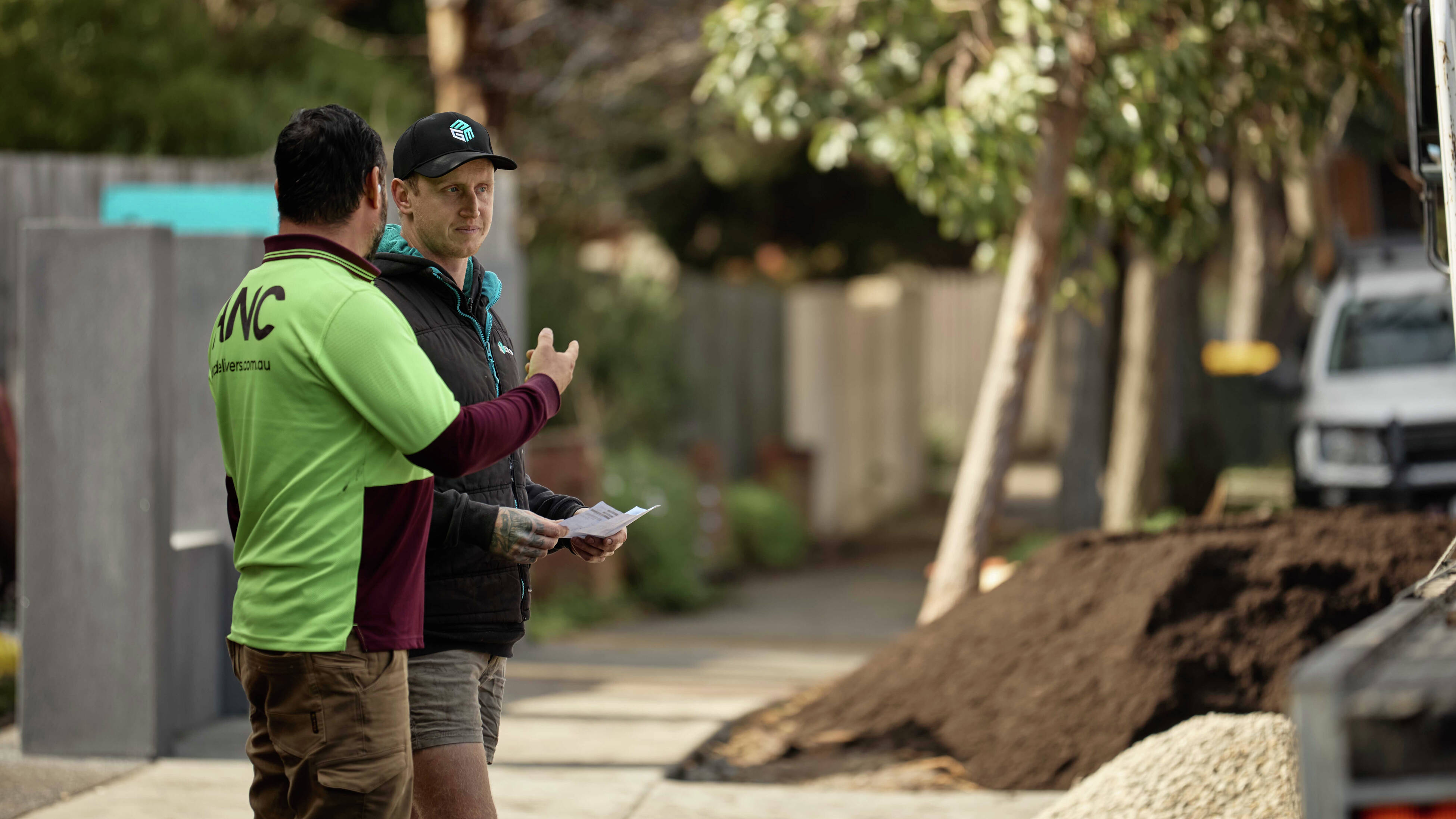 Two men talking outdoors in a residential street, with a pile of soil nearby