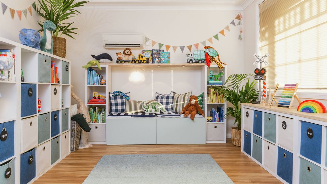 Bright, organized playroom with blue-and-white storage cubbies, toys on shelves, a light rug, and large windows letting in natural light.