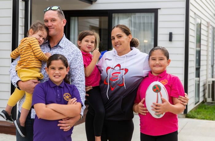 A group of adults and children stand together outside a modern white house. One adult holds a small child, while another adult stands beside several children. One child in a purple shirt stands with arms crossed, and another child in a bright pink shirt holds a rugby ball. The group appears to be posing for a family or community photo.