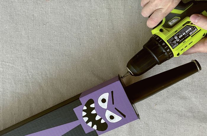 Hands using a drill to screw a furniture foot to a black block decorated with a purple witch face.