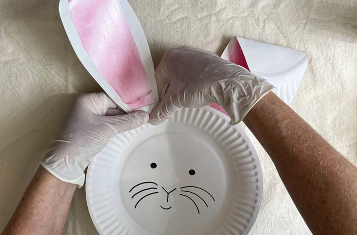 Gloved hands glue paper bunny ears onto a paper plate with a bunny face drawn on it