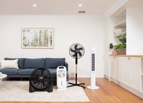 Range of cooling appliances including a floor fan, evaporative cooler, tower fan and air circulator displayed in a modern living room with a blue sofa and timber flooring.