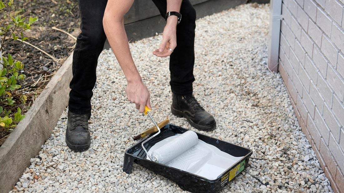 A person standing by a brick wall rolling a paint roller in a tray of white paint