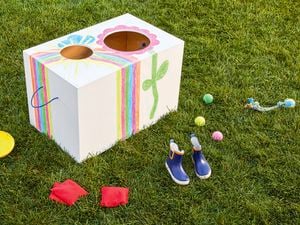 A white wooden toy box decorated with colourful drawings sitting on grass, surrounded by toys and gumboots