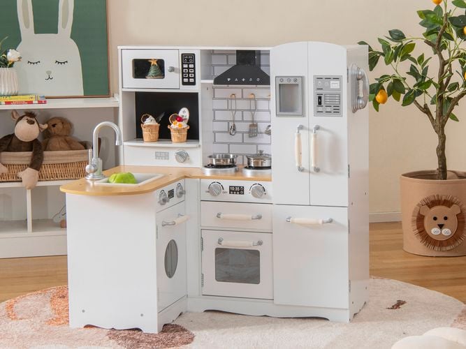A white kids kitchen playset on a rug, with a pot plant and shelving unit next to it in a room.