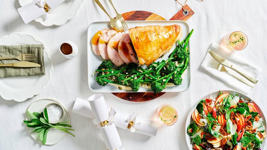 Image of a main dish of Turkey Roulade with broccolini and beans with a zesty dressing, a side dish of fresh salad on a white linen table setting with cutlery and drinks.