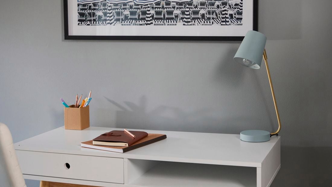 Painted metal desk lamp sits on a desk in front of a grey wall.
