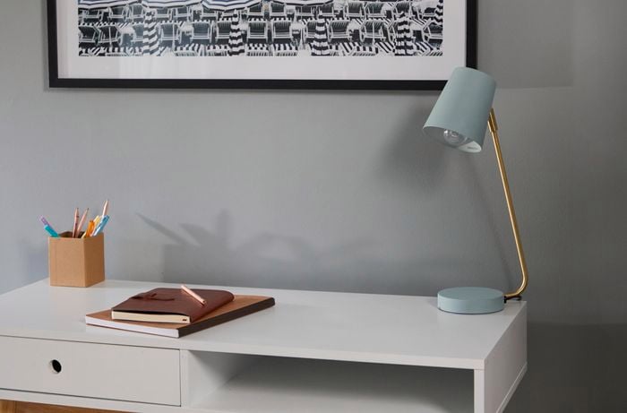 Painted metal desk lamp sits on a desk in front of a grey wall.