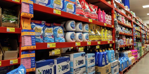 Cleaning supplies aisle in a hardware warehouse, featuring red shelving stocked with laundry detergents, cleaning products, gloves, mop-up cloths and bulk-sized containers. Products are neatly arranged with price labels displayed along the shelves.