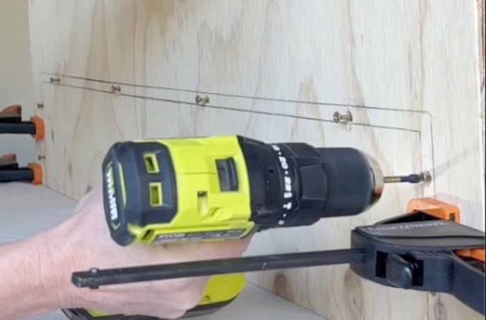 Plywood positioned upright on bench with hands using a drill to make holes in wood.