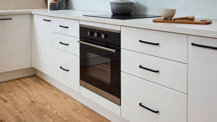 Modern white kitchen cabinetry with black handles, built-in oven and stovetop, light wood flooring, and minimalist benchtop styling with cooking utensils and decor.
