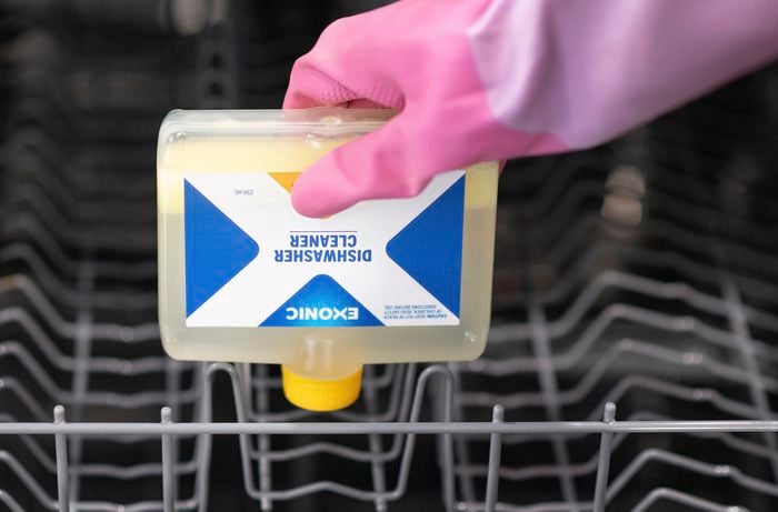 A hand in pink gloves is holding an exonic dishwasher cleaner positioning it downwards towards the dishwasher rack.