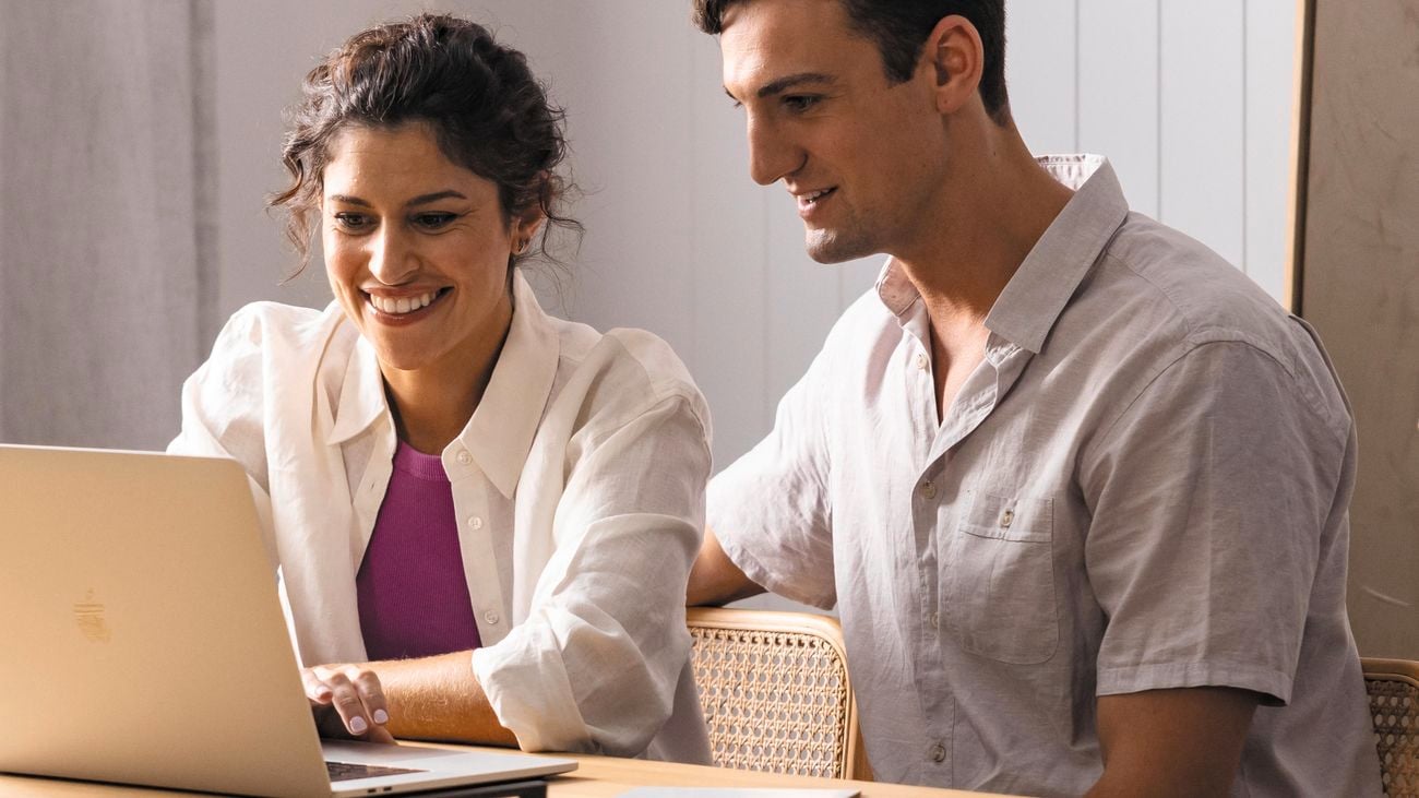 Man and woman sitting at wooden dining table looking at laptop.