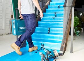 Construction site interior showing protective floor covering installed over stairs and flooring, with power tools and hearing protection placed nearby and a person carrying a tool case up the stairs.