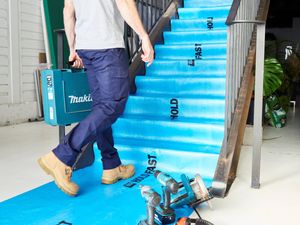 Construction site interior showing protective floor covering installed over stairs and flooring, with power tools and hearing protection placed nearby and a person carrying a tool case up the stairs.
