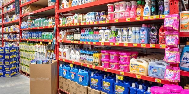 Wide view of a Bunnings retail aisle with red industrial shelving stocked with cleaning and laundry products, including detergents, stain removers, disinfectants, wipes and fabric softeners. Yellow price labels line the shelves, and cartons and bulk containers are stored on lower shelves and pallets.