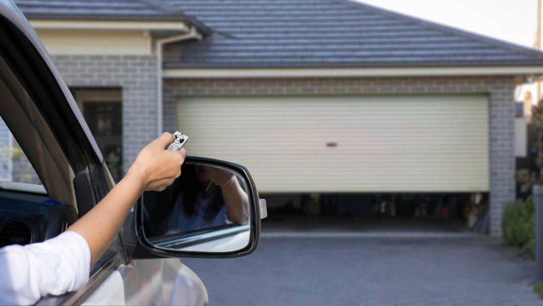 A driver with his/her hand out of the car window to open a garage door using a remote.