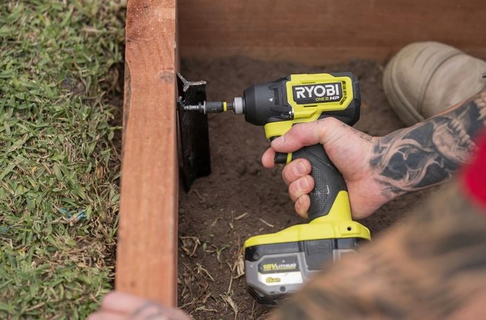 A person is using a Ryobi power drill to fasten a screw into a steel post, securing a wooden sleeper frame. 