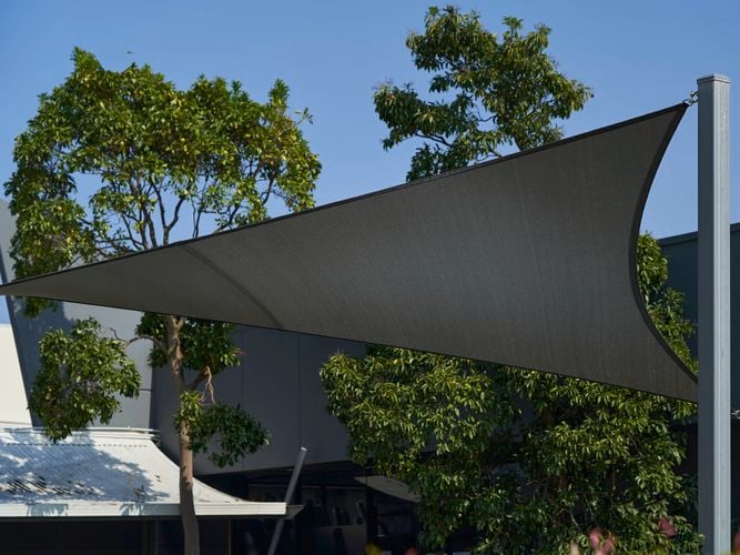 Black triangular shade sail installed outdoors between three metal poles, with trees and a building in the background under a clear blue sky.