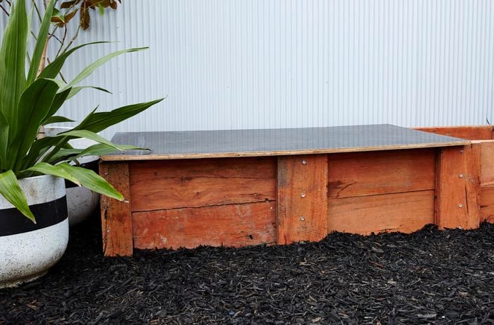 A completed sleeper compost bin sitting amongst dark tanbark in front of a corrugated iron wall