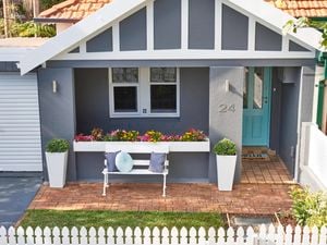 Grey house exterior with colourful potted plants, chair with cushions and a blue front door.