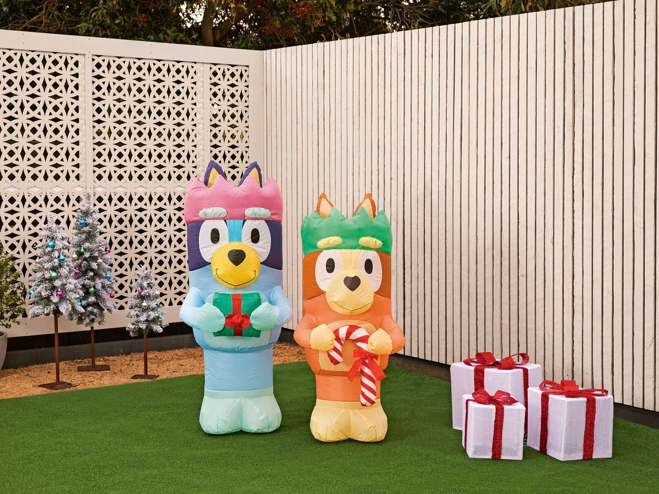 Two inflatable Bluey characters with Christmas gifts and candy canes on a lawn, next to a small tree and wrapped presents against a white fence.