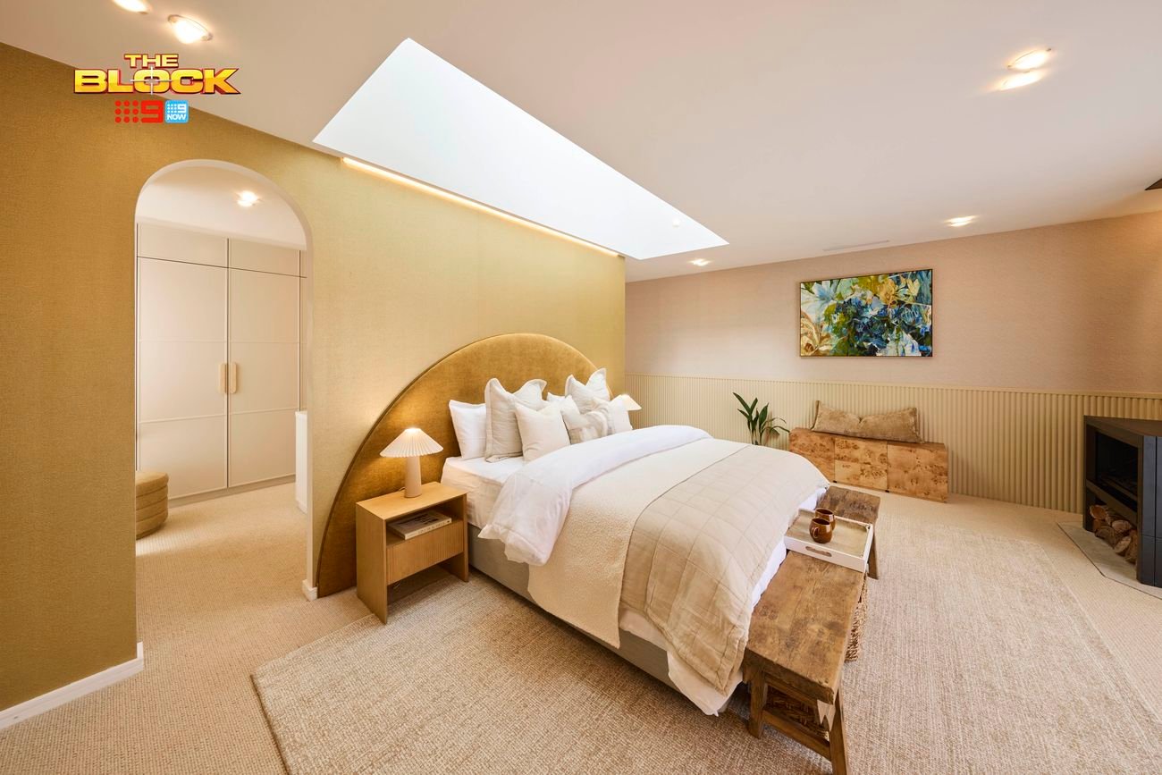 A tastefully decorated bedroom with a large bed centered under a skylight, dressed in a beige blanket and multiple pillows. Two bedside tables with lamps flank the bed. A fireplace is visible on the right, with a bench at the foot of the bed. A colorful painting hangs above a small plant and a piece of furniture. The room features neutral-toned walls and carpeted flooring.