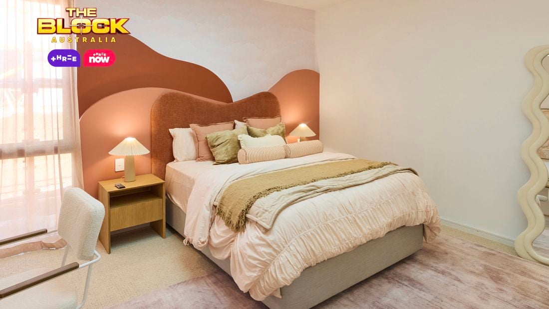Cream bedroom with a queen bed, desk and mirror on a blush rug and a red and orange hill style bedhead.