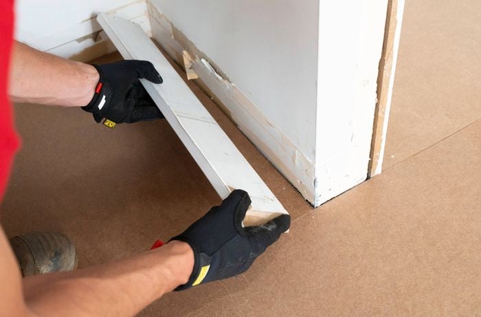 A person in black gloves is holding a skirting board near the bottom of a wall corner.