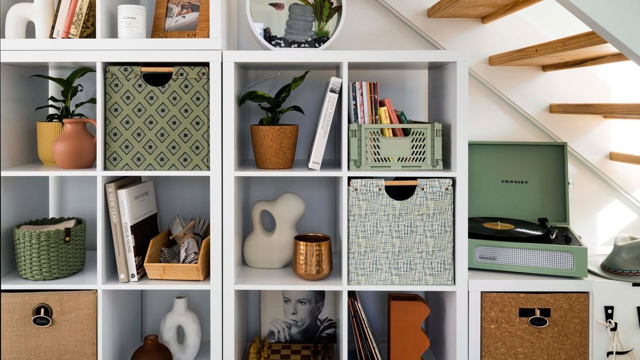 Shelving units underneath stairs with baskets in it, fish bowl on it, plants in it and a record player on it