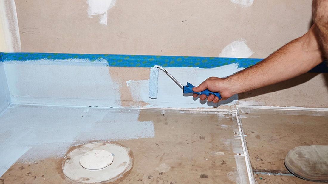A person using a roller to apply waterproofing agent on a wall up to a masking tape line
