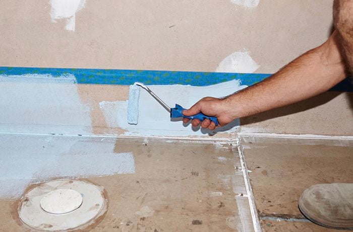 A person using a roller to apply waterproofing agent on a wall up to a masking tape line