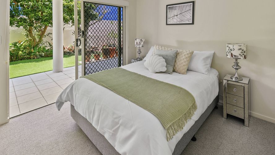 Bright bedroom featuring a queen-size bed with neutral furnishings, bedside tables and lamps, and sliding door access to a covered patio and landscaped garden.
