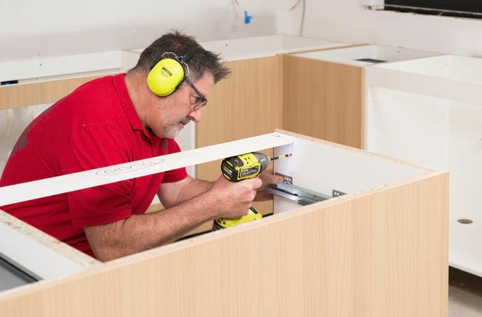 A Bunnings Team Member wearing ear protection is using a power drill to attach an end panel to the exposed side of the final cabinet.