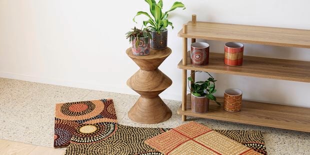 A couple of coloured small rugs on a larger speckled white rug next to a pot stand with plants on it and a shelving unit with plants on it.