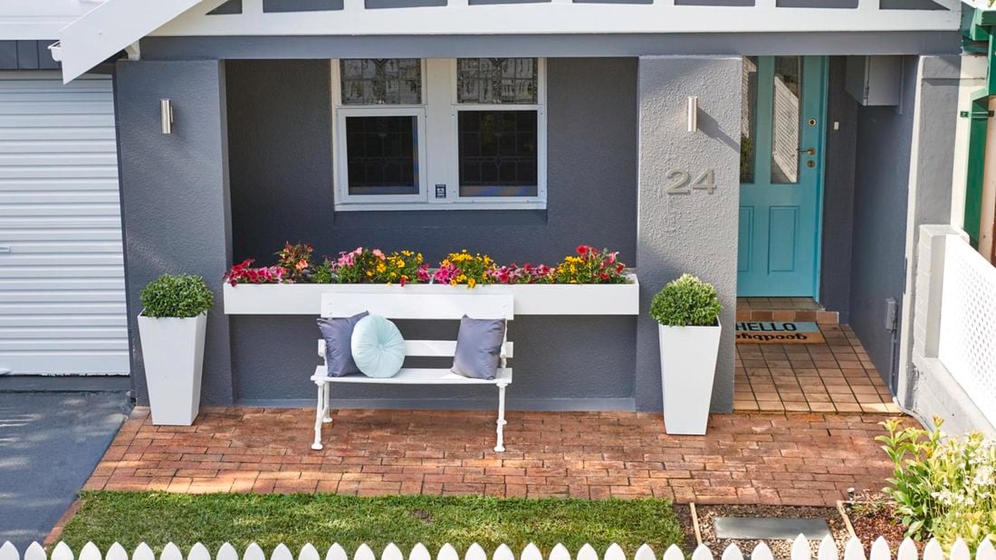 Grey house exterior with colourful potted plants, chair with cushions and a blue front door.