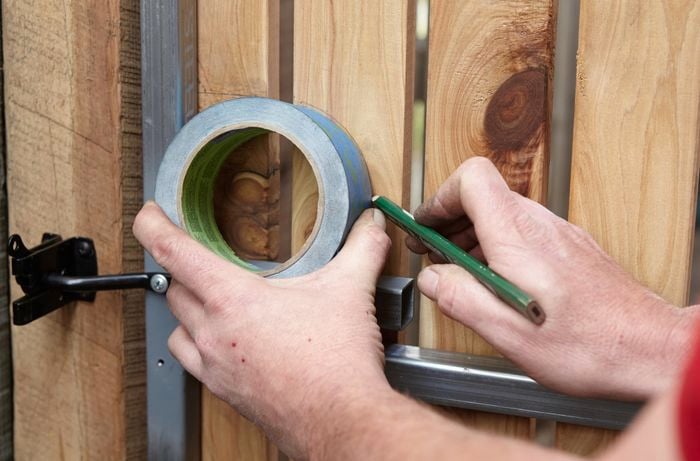 Person holding roll of tape against fence and drawing an outline with a pencil.