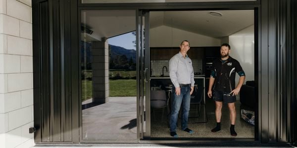 Two people stand inside a modern home with large sliding glass doors open to a covered patio and a scenic outdoor landscape.